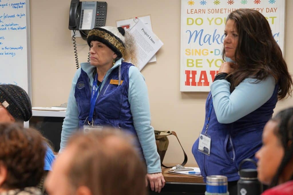 Two women standing against a wall listening to someone speaking